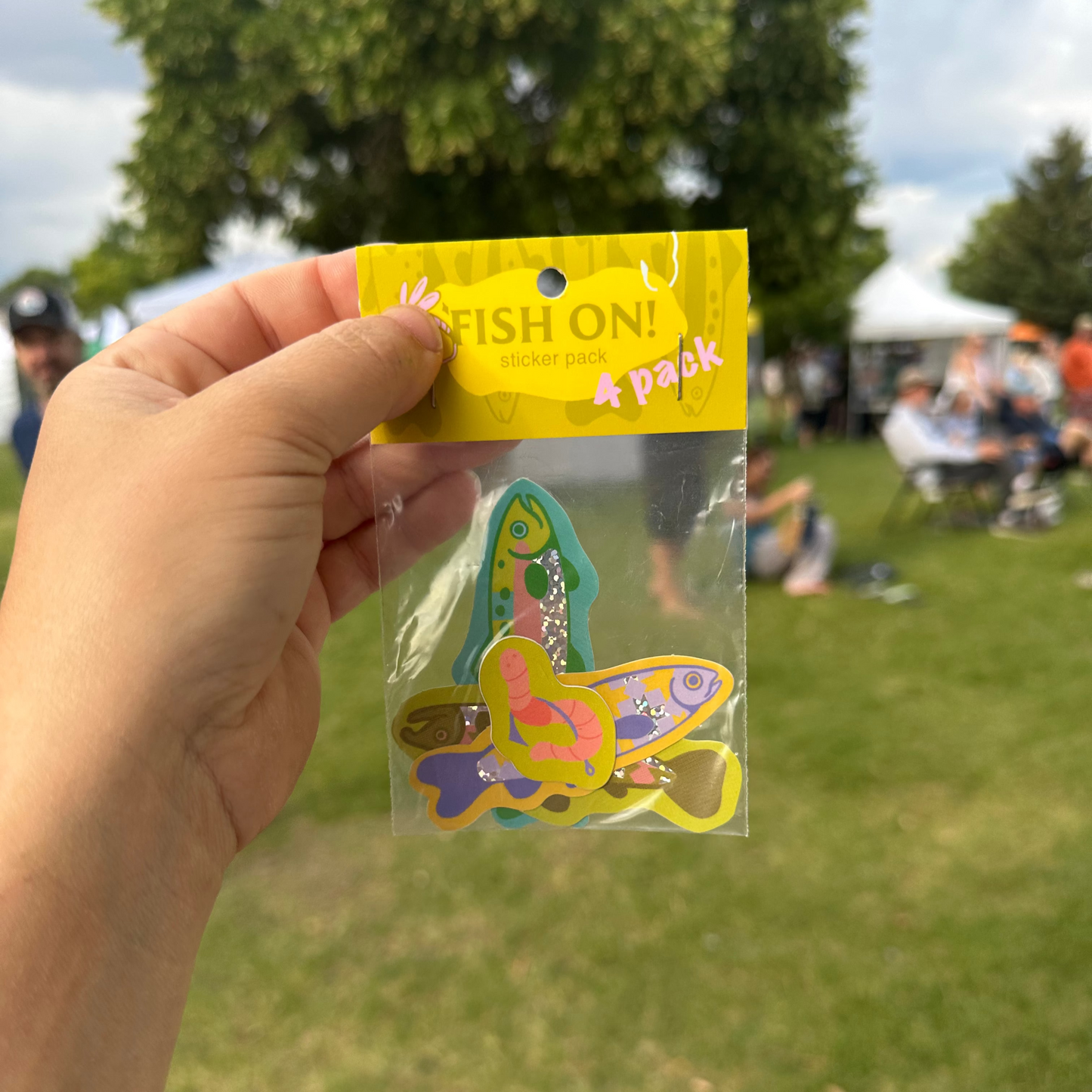 Hand holding a package of colorful fish-shaped earrings with 'FISH ON!' text against a blurred outdoor background.